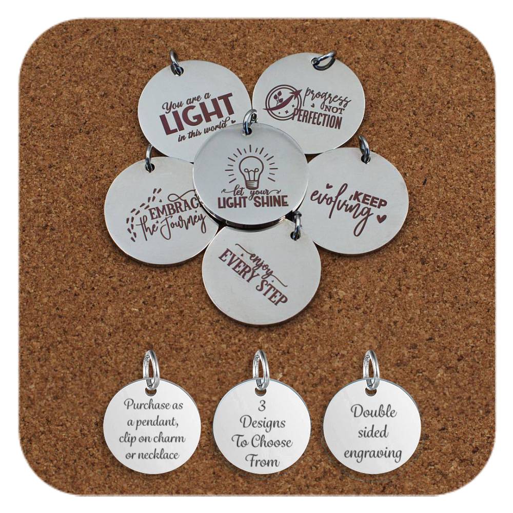 Motivational stainless steel pendants engraved with uplifting phrases such as 'Let Your Light Shine', 'Keep Evolving', and 'Embrace the Journey' displayed on a cork background. Below the pendants are three additional discs showing engraved text that reads: 'Purchase as a pendant, clip on charm or necklace,' '3 Designs To Choose From,' and 'Double sided engraving'