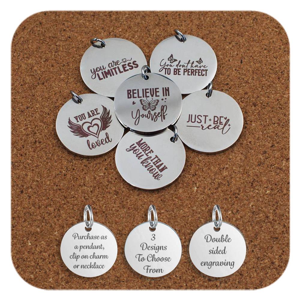 Inspirational stainless steel pendants engraved with messages like 'Believe in Yourself', 'You Are Loved', and 'Just Be Real' arranged on a cork background. Below the pendants are three additional discs showing engraved text that reads: 'Purchase as a pendant, clip on charm or necklace,' '3 Designs To Choose From,' and 'Double sided engraving'
