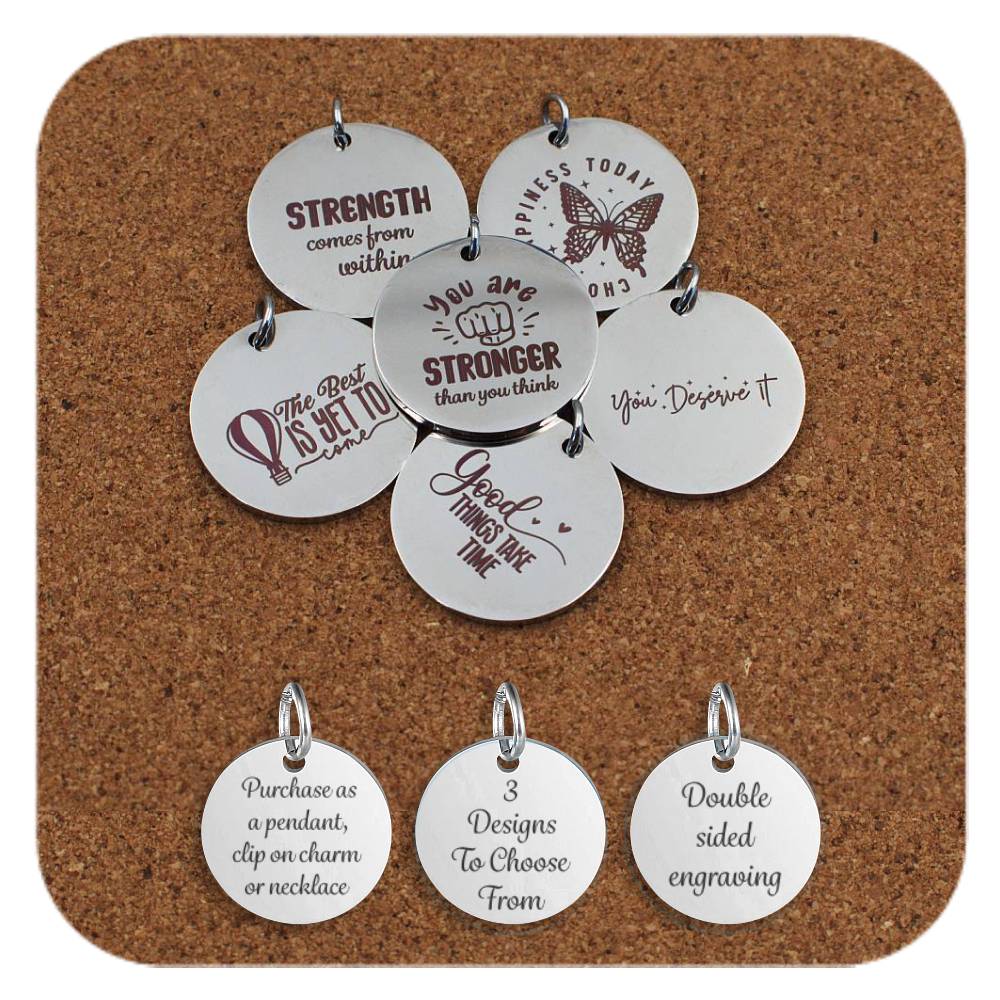 Round stainless steel pendants engraved with uplifting messages such as 'You Are Stronger Than You Think', 'Strength Comes from Within', and 'The Best Is Yet to Come' displayed on a cork background. Below the pendants are three additional discs showing engraved text that reads: 'Purchase as a pendant, clip on charm or necklace,' '3 Designs To Choose From,' and 'Double sided engraving'