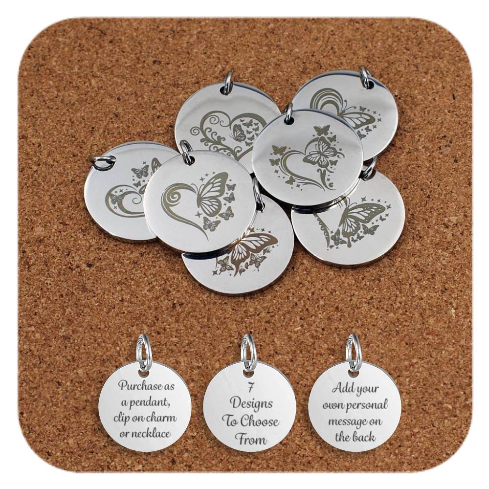 Stainless steel pendants featuring engraved designs of butterflies and heart motifs, with decorative swirls and stars, displayed on a cork background. Below the pendants are three additional discs showing engraved text that reads: 'Purchase as a pendant, clip on charm or necklace,' '7 Designs To Choose From,' and 'Add your own personal message on the back'