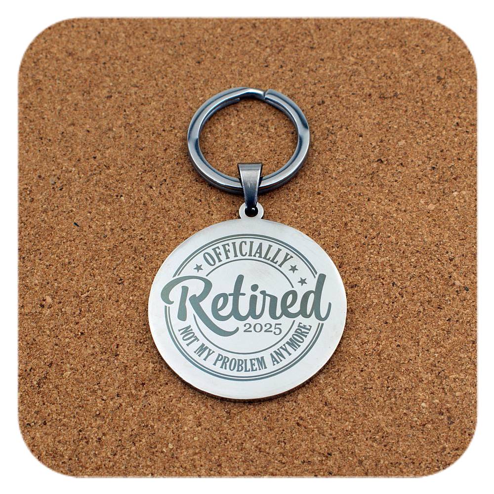 Silver keychain with 'Officially Retired 2025 Not My Problem Anymore' text on a cork background