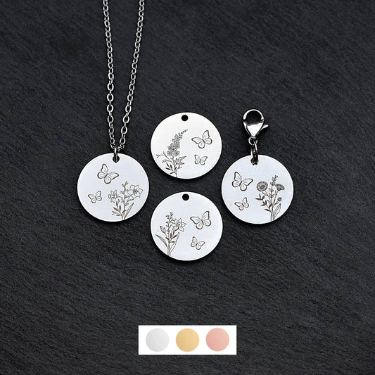 Four silver round disc pendants on a black slate background showing four different birth flower and butterfly designs. One has a silver necklace attached by a jump ring and another has a lobster clasp and split ring attached to it