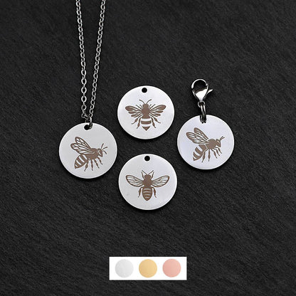 Four silver round disc pendants on a black slate background showing four different bee designs. One has a silver necklace attached by a jump ring and another has a lobster clasp and split ring attached to it