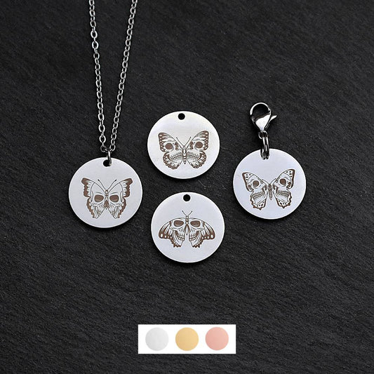 Four silver round disc pendants on a black slate background showing four different skull butterfly designs. One has a silver necklace attached by a jump ring and another has a lobster clasp and split ring attached to it