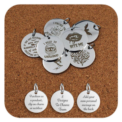 A selection of stainless steel pendants engraved with playful Halloween-themed designs and phrases, including vampires, bats, mummies, and humorous spooky quotes, displayed on a cork background. Below the pendants are three additional discs showing engraved text that reads: 'Purchase as a pendant, clip on charm or necklace,' '7 Designs To Choose From,' and 'Add your own personal message on the back'