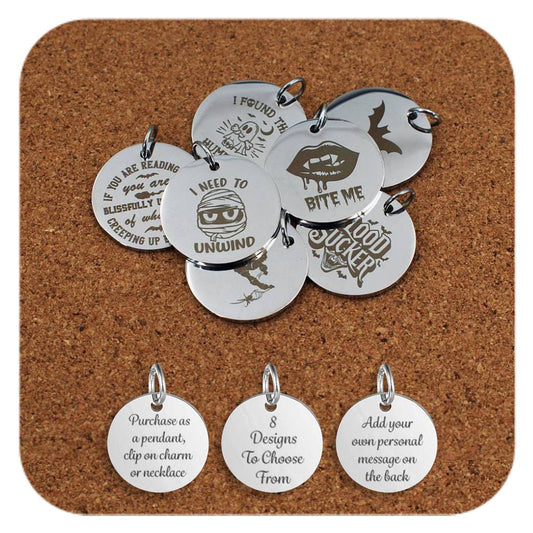 A selection of stainless steel pendants engraved with playful Halloween-themed designs and phrases, including vampires, bats, mummies, and humorous spooky quotes, displayed on a cork background. Below the pendants are three additional discs showing engraved text that reads: 'Purchase as a pendant, clip on charm or necklace,' '7 Designs To Choose From,' and 'Add your own personal message on the back'