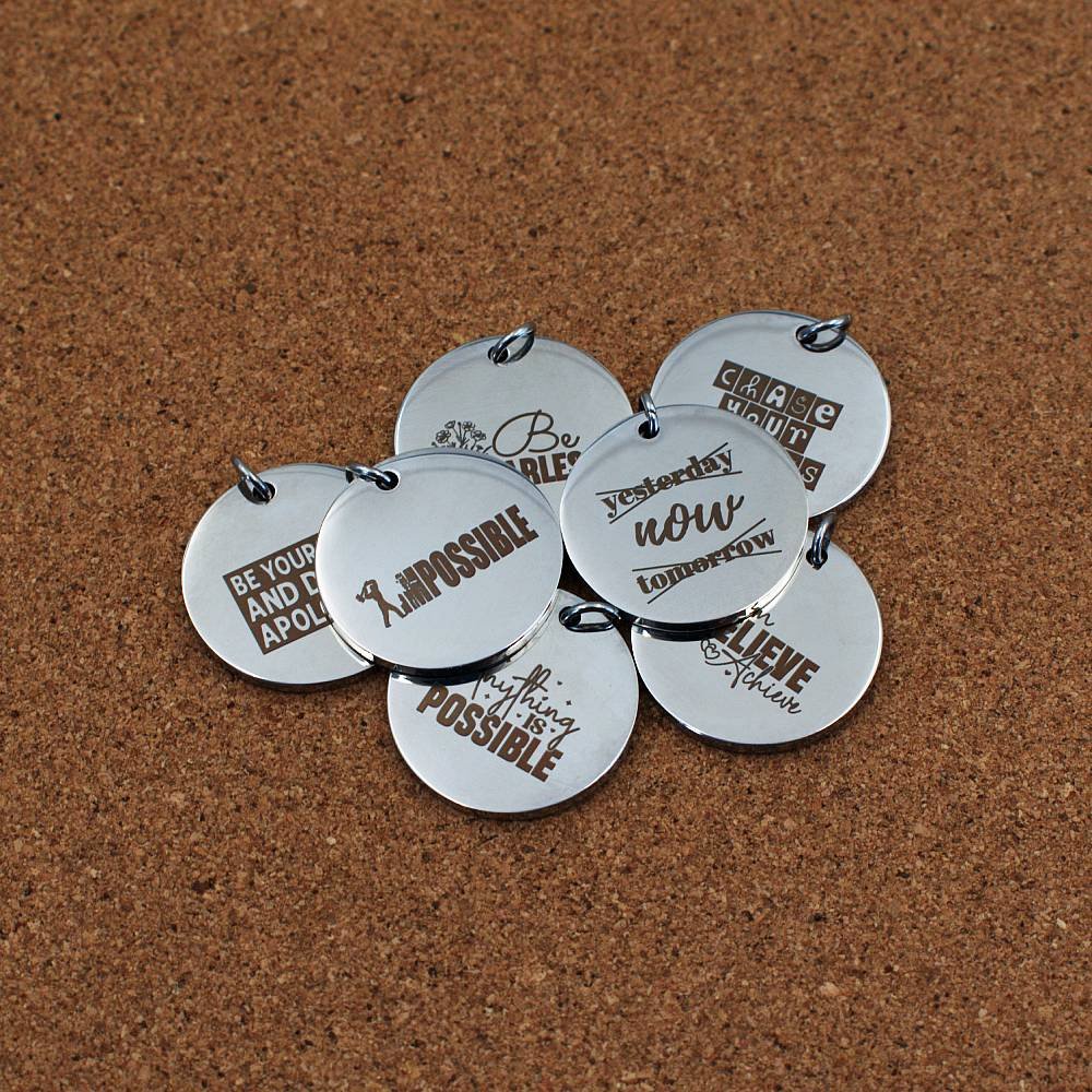 Round stainless steel pendants engraved with motivational quotes such as 'Be You and Don’t Apologise,' 'Chase Your Goals,' 'Believe & Achieve,' and 'Nothing is Impossible.' Laid out on a cork background in a casual arrangement