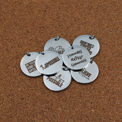 Round stainless steel pendants engraved with motivational quotes such as 'Be You and Don’t Apologise,' 'Chase Your Goals,' 'Believe & Achieve,' and 'Nothing is Impossible.' Laid out on a cork background in a casual arrangement
