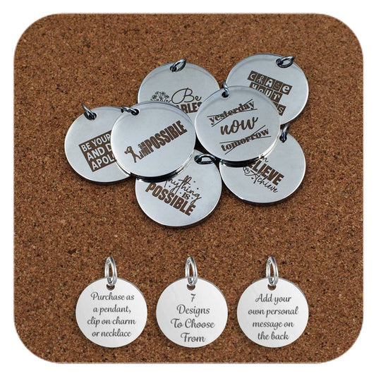 Selection of round stainless steel pendants engraved with motivational quotes, including ‘Be Fearless,’ ‘Chase Your Goals,’ and ‘Impossible.’ displayed on a cork background. Below the pendants are three additional discs showing engraved text that reads: 'Purchase as a pendant, clip on charm or necklace,' '7 Designs To Choose From,' and 'Add your own personal message on the back'