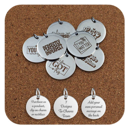Engraved stainless steel pendants with uplifting messages like ‘Perfectly Imperfect,’ ‘You Matter,’ and ‘You Got This.’ displayed on a corkboard. Below the pendants are three additional discs showing engraved text that reads: 'Purchase as a pendant, clip on charm or necklace,' '7 Designs To Choose From,' and 'Add your own personal message on the back'