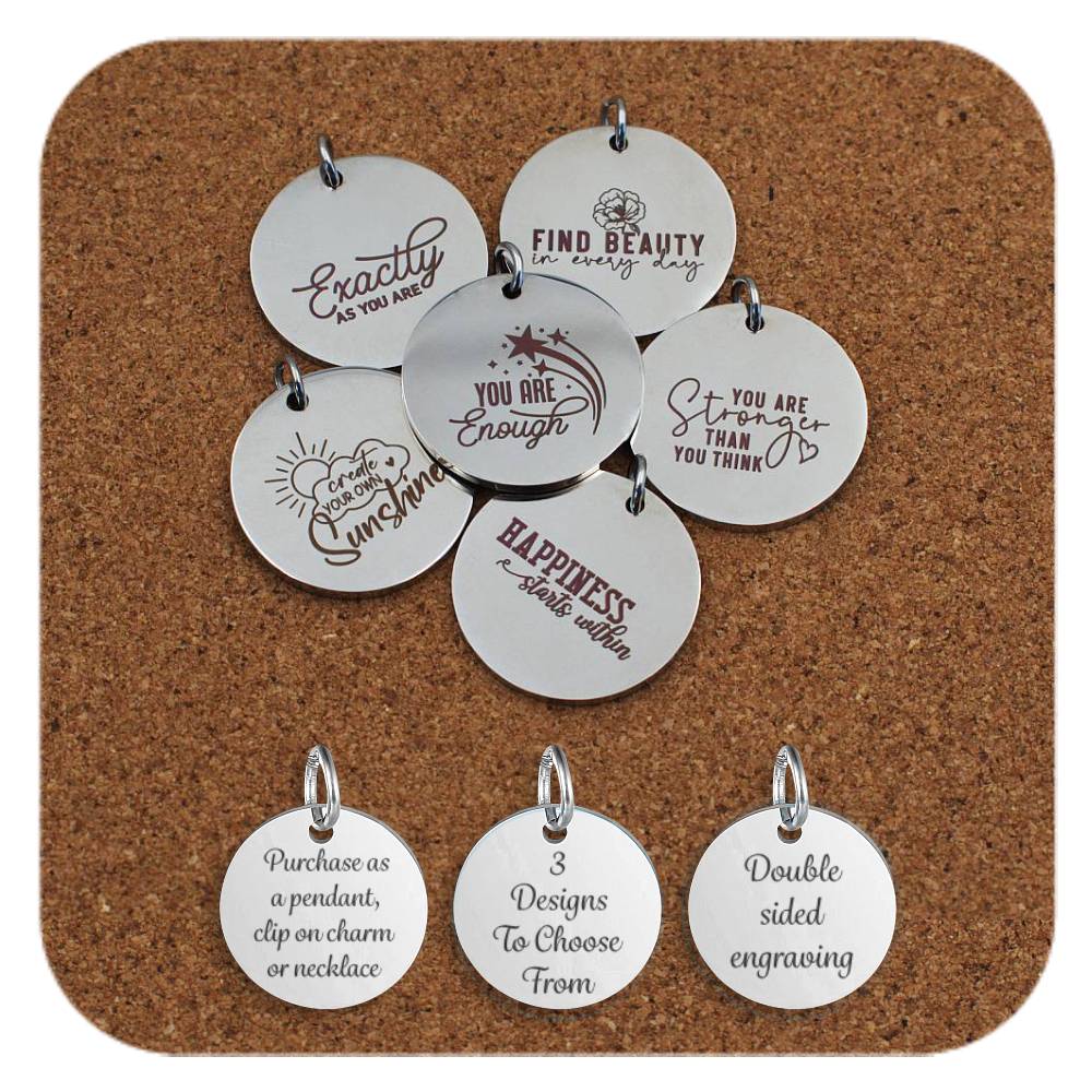 Collection of round stainless steel pendants engraved with affirmations like 'You Are Enough', 'You Are Stronger Than You Think', and 'Create Your Own Sunshine' displayed on a cork background. Below the pendants are three additional discs showing engraved text that reads: 'Purchase as a pendant, clip on charm or necklace,' '3 Designs To Choose From,' and 'Double sided engraving'