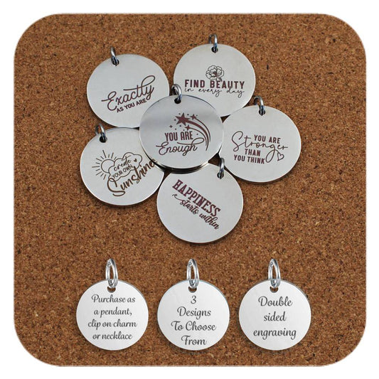 Collection of round stainless steel pendants engraved with affirmations like 'You Are Enough', 'You Are Stronger Than You Think', and 'Create Your Own Sunshine' displayed on a cork background. Below the pendants are three additional discs showing engraved text that reads: 'Purchase as a pendant, clip on charm or necklace,' '3 Designs To Choose From,' and 'Double sided engraving'