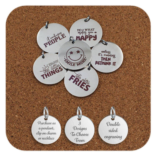 Round stainless steel pendants engraved with light-hearted and motivational quotes including 'Do what makes you happy', 'Smile more', 'Enjoy the little things', and 'I like the extra fries'. Displayed on a cork background with three informational charms at the bottom reading: 'Purchase as a pendant, clip on charm or necklace', '3 Designs To Choose From', and 'Double sided engraving'.