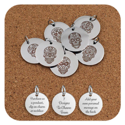 A group of stainless steel pendants engraved with decorative sugar skull designs, inspired by Day of the Dead motifs, displayed on a cork background. Below the pendants are three additional discs showing engraved text that reads: 'Purchase as a pendant, clip on charm or necklace,' '7 Designs To Choose From,' and 'Add your own personal message on the back'