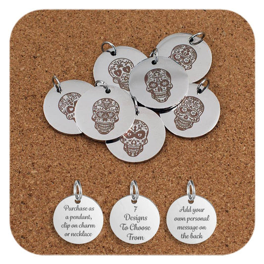 A group of stainless steel pendants engraved with decorative sugar skull designs, inspired by Day of the Dead motifs, displayed on a cork background. Below the pendants are three additional discs showing engraved text that reads: 'Purchase as a pendant, clip on charm or necklace,' '7 Designs To Choose From,' and 'Add your own personal message on the back'