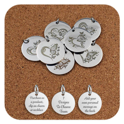Stainless steel pendants featuring engraved designs of butterflies and heart motifs, with decorative swirls and stars, displayed on a cork background. Below the pendants are three additional discs showing engraved text that reads: 'Purchase as a pendant, clip on charm or necklace,' '7 Designs To Choose From,' and 'Add your own personal message on the back'