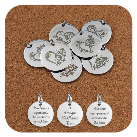 Stainless steel pendants featuring engraved designs of butterflies and heart motifs, with decorative swirls and stars, displayed on a cork background. Below the pendants are three additional discs showing engraved text that reads: 'Purchase as a pendant, clip on charm or necklace,' '7 Designs To Choose From,' and 'Add your own personal message on the back'
