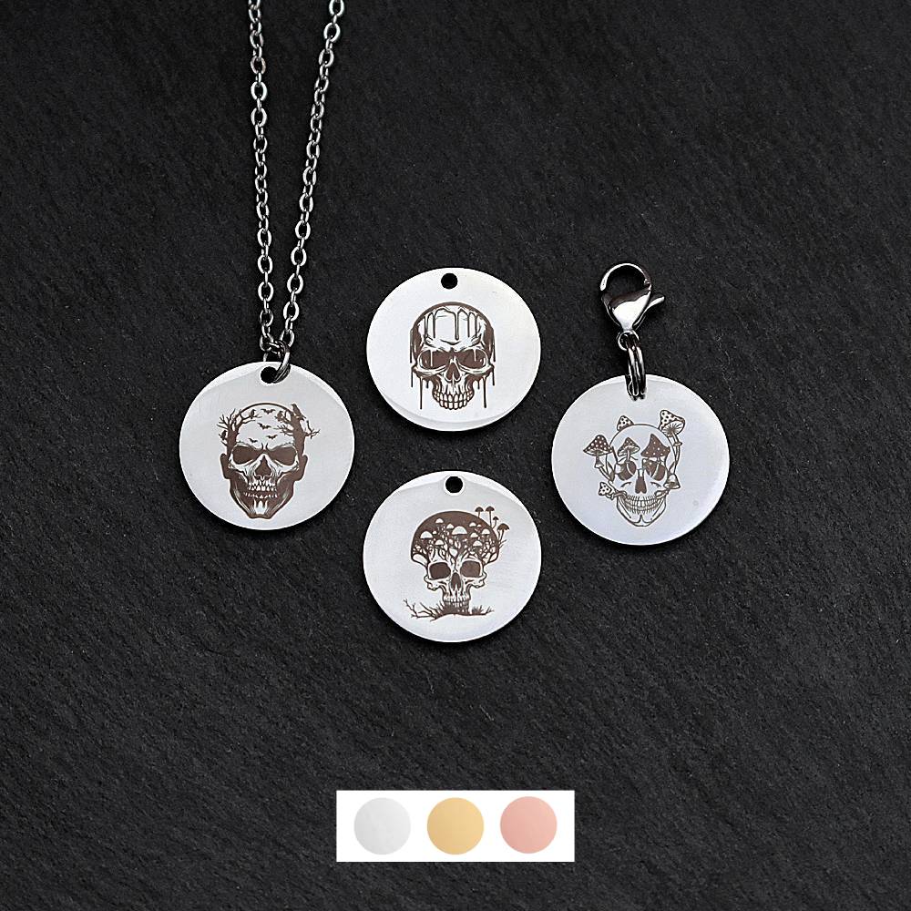 Four silver round disc pendants on a black slate background showing four different creepy skull designs. One has a silver necklace attached by a jump ring and another has a lobster clasp and split ring attached to it