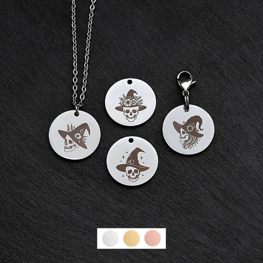 Four silver round disc pendants on a black slate background showing four different skulls wearing a witch hat designs. One has a silver necklace attached by a jump ring and another has a lobster clasp and split ring attached to it