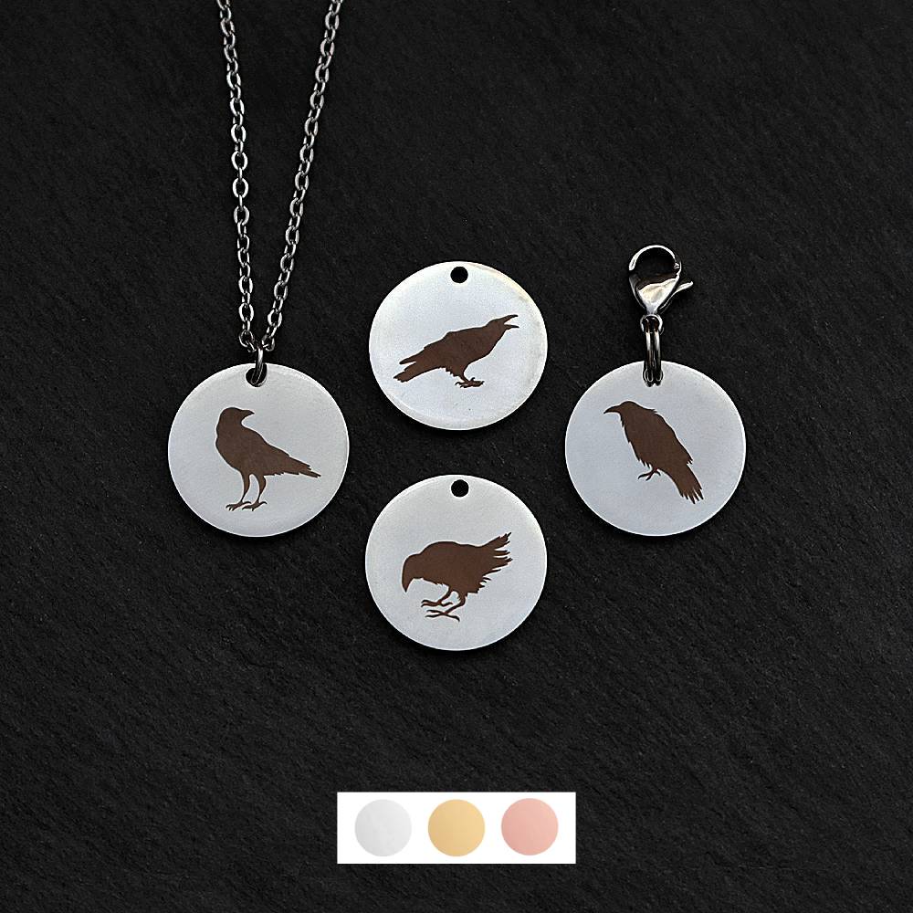 Four silver round disc pendants on a black slate background showing four different black raven rook birds. One has a silver necklace attached by a jump ring and another has a lobster clasp and split ring attached to it