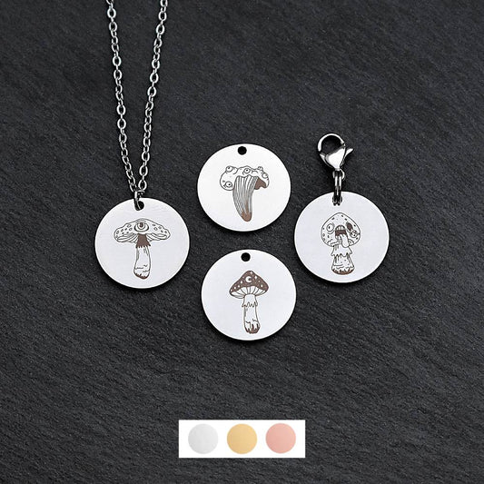 Four silver round disc pendants on a black slate background showing four different creepy mushroom toadstool designs. One has a silver necklace attached by a jump ring and another has a lobster clasp and split ring attached to it