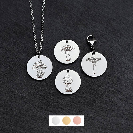 Four silver round disc pendants on a black slate background showing four different creepy mushroom toadstool designs. One has a silver necklace attached by a jump ring and another has a lobster clasp and split ring attached to it