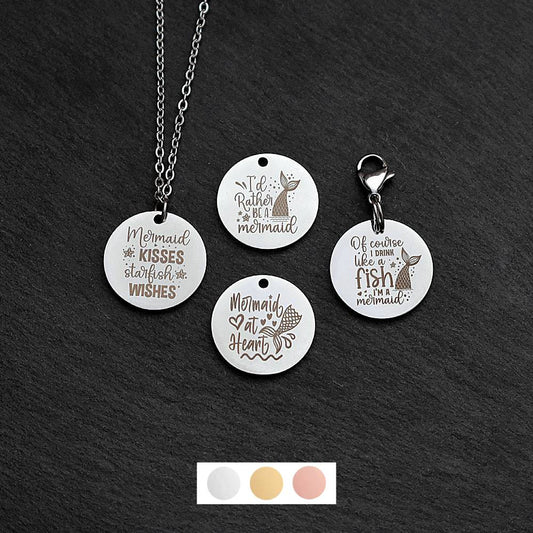 Four silver round disc pendants on a black slate background showing four different mermaid quote designs. One has a silver necklace attached by a jump ring and another has a lobster clasp and split ring attached to it